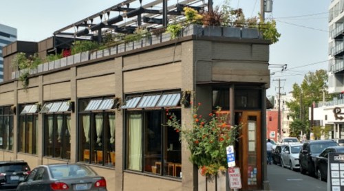A modern restaurant with large windows and a rooftop garden, located on a city street.