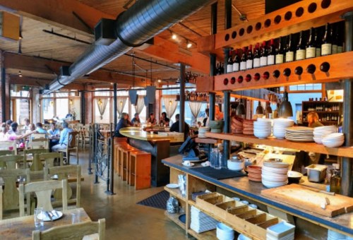 A cozy restaurant interior with wooden decor, tables filled with diners, and a bar displaying wine bottles.