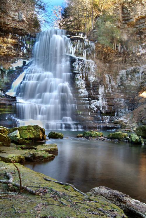 A serene waterfall cascades over rocky cliffs, surrounded by icicles and lush greenery, reflecting in a calm pool below.