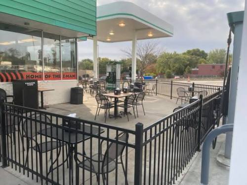 Outdoor seating area with tables and chairs at a gas station, surrounded by a black fence and green building.