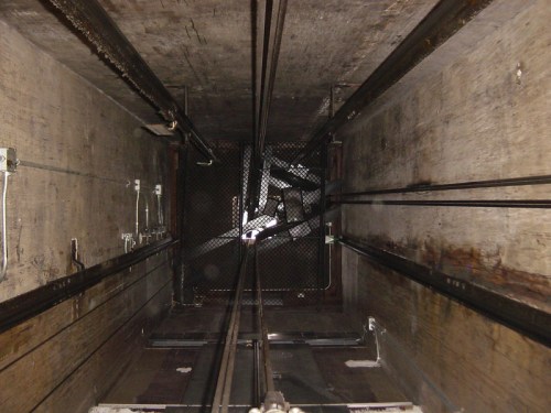 View looking up an elevator shaft, featuring metal cables and a grid-like ceiling. Walls are concrete and weathered.