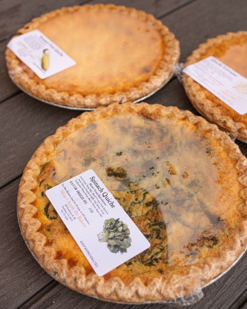 Three spinach quiches in pie tins, each covered with plastic wrap and labeled with ingredients and cooking instructions.