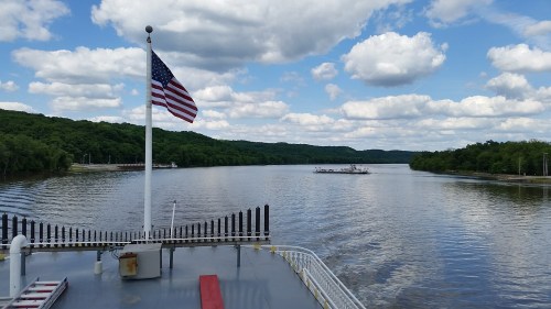 View from a boat on a river, featuring a flag, lush green hills, and a cloudy blue sky.