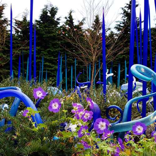 Colorful glass sculptures rise among vibrant flowers and greenery in a lush garden setting.