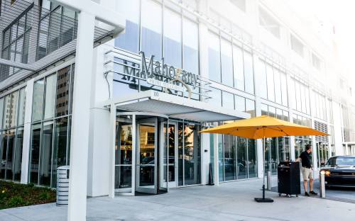 Entrance of a modern building with large glass windows and a yellow umbrella outside. A person is near a cart.