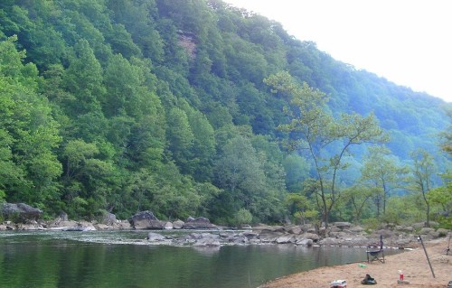 A serene riverbank surrounded by lush green trees and rocky formations, with a calm water surface reflecting the landscape.