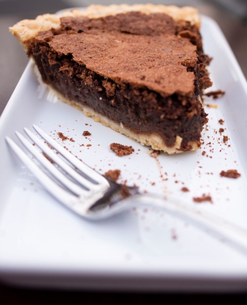 A slice of rich chocolate pie on a white plate, with a fork and crumbs scattered around.