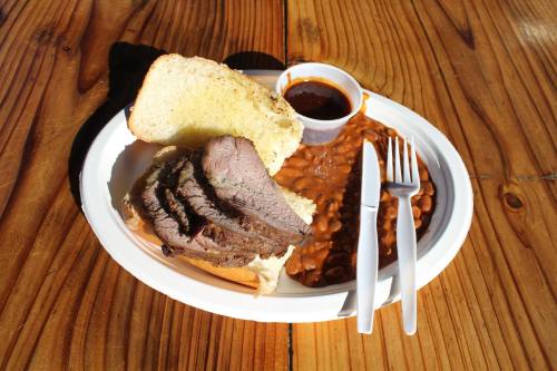 A plate with sliced beef on bread, baked beans, and a small cup of sauce, set on a wooden table.