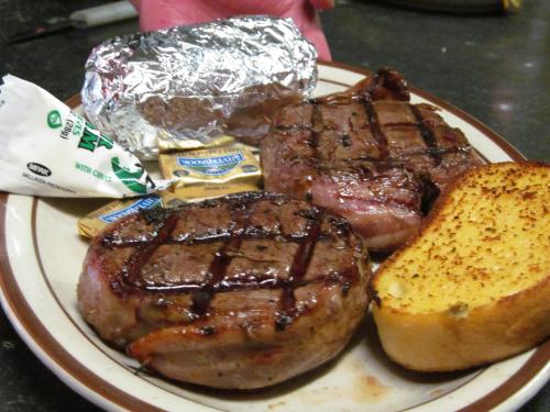A plate with two grilled steaks, a baked potato, butter packets, and a slice of toasted bread.