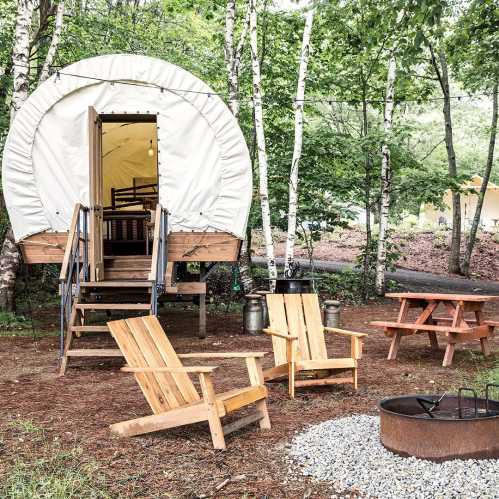 A cozy glamping tent on stilts surrounded by trees, with wooden chairs, a picnic table, and a fire pit nearby.