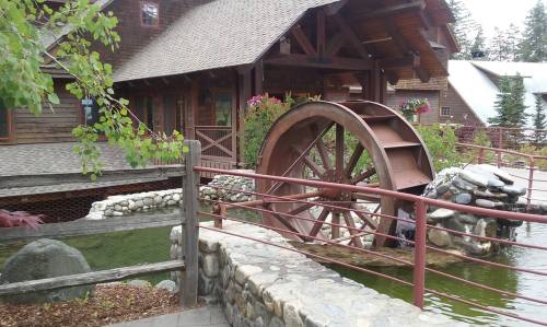 A wooden water wheel beside a stone bridge, with a rustic cabin and greenery in the background.