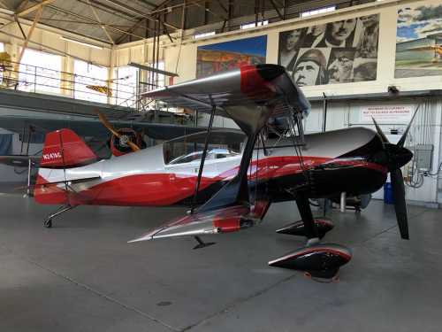 A sleek red and silver biplane parked inside a hangar, with vintage aviation photos on the wall behind it.