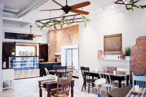 A cozy café interior with wooden tables, chairs, exposed brick walls, and ceiling fans, featuring a display fridge.