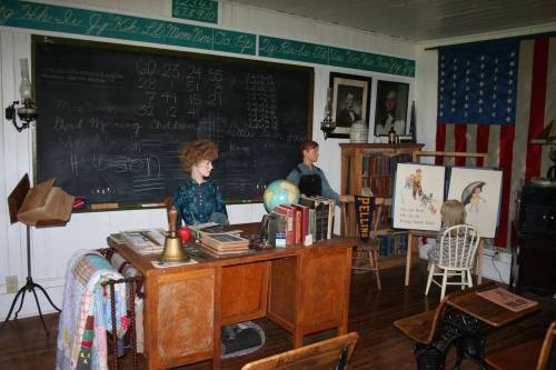 A vintage classroom scene with mannequins at desks, a chalkboard, books, and an American flag in the background.