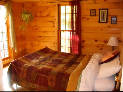 Cozy wooden bedroom with a bed, colorful quilt, and large windows letting in natural light.