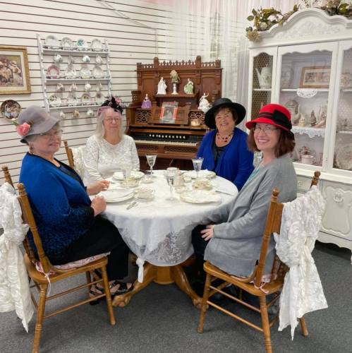 Four women in hats sit at a table set for tea, surrounded by vintage decor and china in a cozy room.