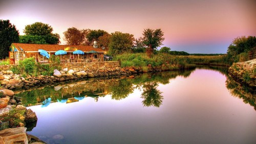 A tranquil river scene with a rustic building, lush greenery, and reflections at sunset.