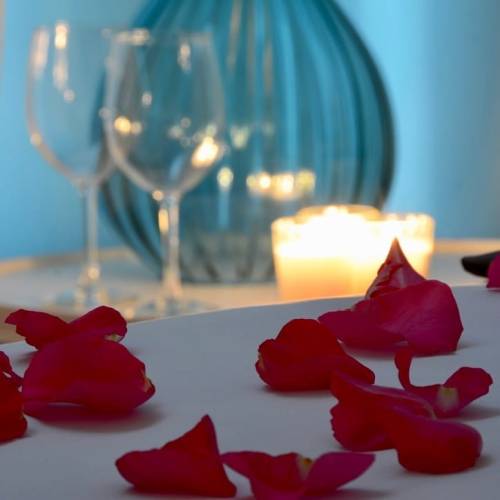 A romantic table setting with rose petals, two glasses, a blue vase, and candles glowing softly in the background.