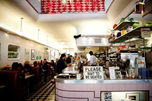 A bustling diner interior with patrons seated at booths and a busy counter, featuring a "Please wait to be seated" sign.