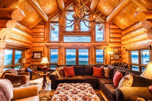 Cozy log cabin interior with large windows, a plush sofa, and scenic mountain views. Warm lighting enhances the rustic charm.