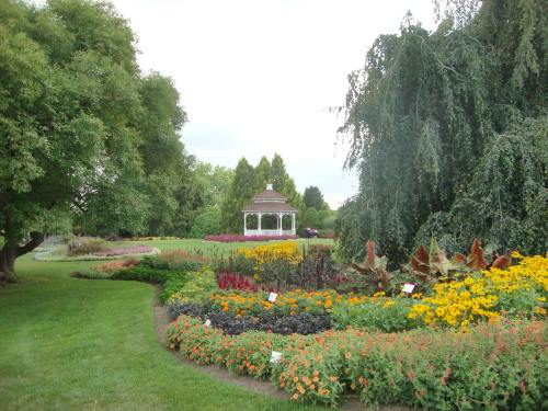A lush garden with colorful flowers and a gazebo in the background, surrounded by trees under a cloudy sky.