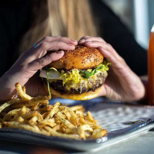 A person holds a juicy burger with lettuce and onion, accompanied by a plate of golden fries.