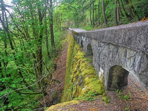 A moss-covered stone bridge alongside a winding road, surrounded by lush green trees and a steep drop-off.