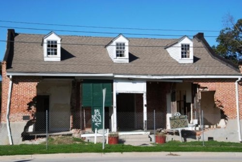 Abandoned building with brick walls, missing sections, and a fenced-off entrance, showing signs of decay and neglect.