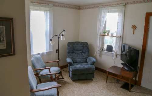 Cozy living room with two chairs, a recliner, a TV, and natural light from windows. Simple decor and warm atmosphere.