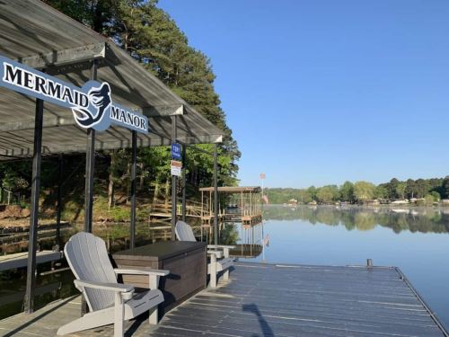 A peaceful lakeside view featuring a dock with "Mermaid Manor" sign and wooden chairs overlooking calm water.