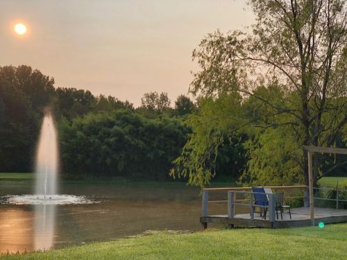 A serene pond with a fountain, surrounded by trees, and a wooden deck with two chairs under a hazy sunset.