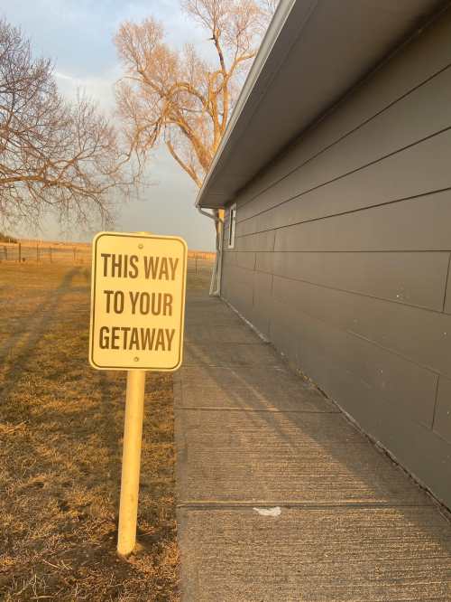 A sign reading "THIS WAY TO YOUR GETAWAY" next to a pathway beside a building, with trees in the background.