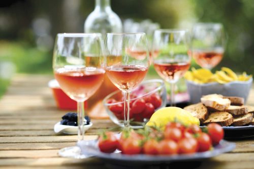 A table set outdoors with glasses of rosé wine, fresh tomatoes, lemon, bread, and snacks, surrounded by greenery.