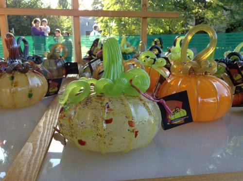 Colorful glass pumpkins with decorative stems displayed on a table, surrounded by greenery and people in the background.