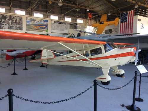 A vintage airplane with an orange stripe displayed in an aviation museum, surrounded by other aircraft and historical images.