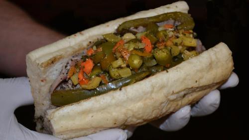 A hand holding a large sandwich filled with sliced meats, pickles, and colorful peppers in a soft bun.