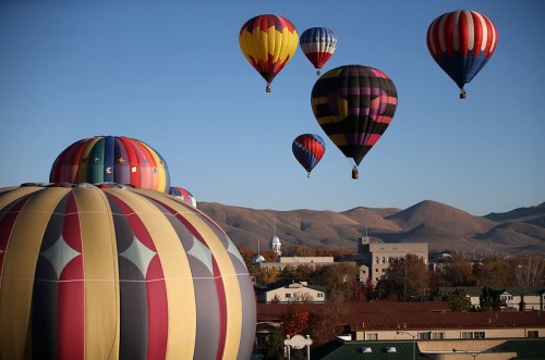 Colorful hot air balloons float over a town with mountains in the background on a clear day.