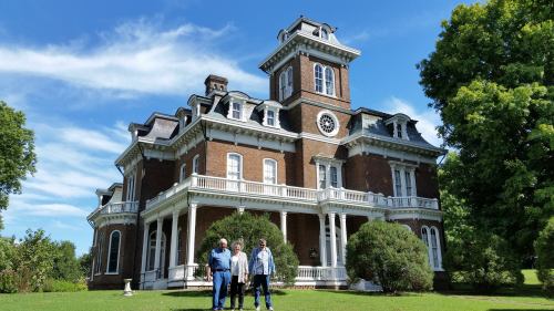 Three people stand in front of a large, historic mansion with a tower, surrounded by greenery and a blue sky.