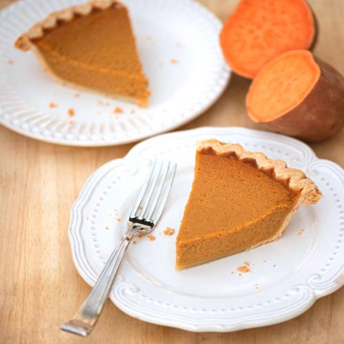 A slice of pumpkin pie on a white plate, with a whole pumpkin and a halved pumpkin beside it on a wooden surface.