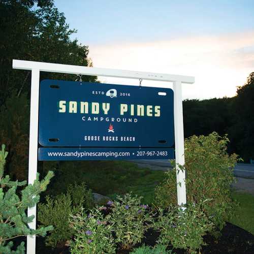 Sign for Sandy Pines Campground, established in 2016, with a scenic background of trees and a sunset.