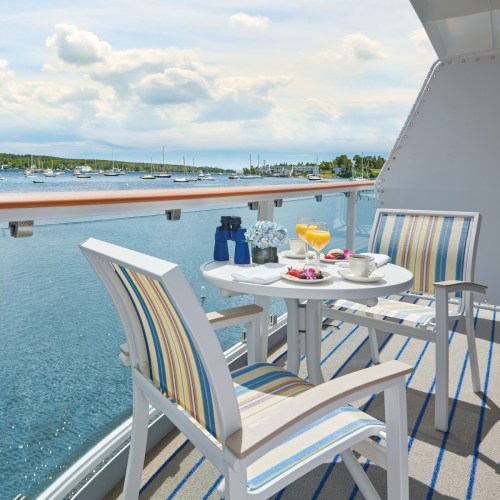 A cozy balcony with striped chairs, a table set with drinks and pastries, overlooking a serene harbor with boats.
