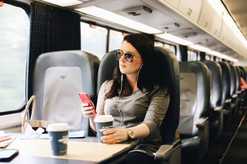A woman in sunglasses sits on a train, holding a phone and sipping coffee, with earbuds in her ears.
