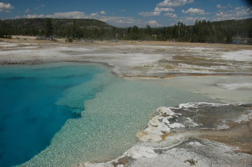 Vibrant blue hot spring surrounded by white mineral deposits and a scenic landscape under a clear sky.