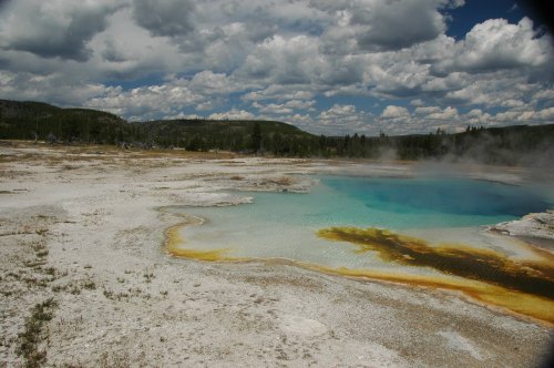 A vibrant geothermal pool with turquoise water and yellow mineral deposits, surrounded by a cloudy sky and forested hills.