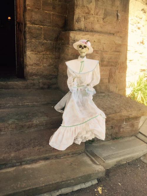 A skeleton dressed in a vintage white dress and hat sits on stone steps, adding a spooky touch to the scene.