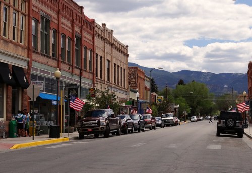 A quaint street lined with brick buildings and parked cars, featuring American flags and mountains in the background.