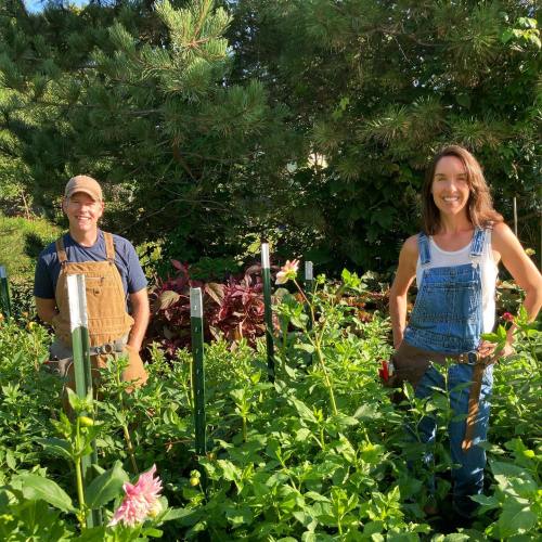Two people in overalls stand among vibrant flowers in a garden, surrounded by greenery and plants.