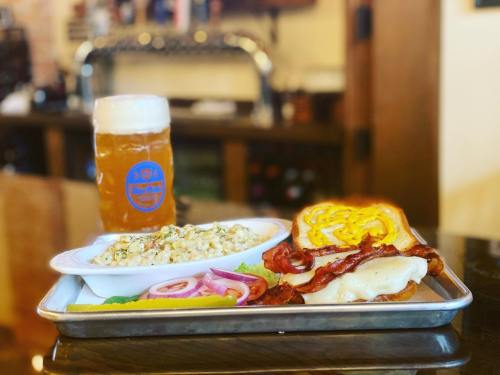 A tray with a sandwich, bacon, macaroni salad, pickles, and a frosty beer in a glass.