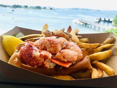 A lobster roll with fries and lemon, set against a scenic waterfront view.