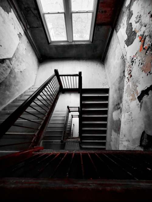 A view looking down a worn staircase with peeling walls and a large window at the top, creating a moody atmosphere.
